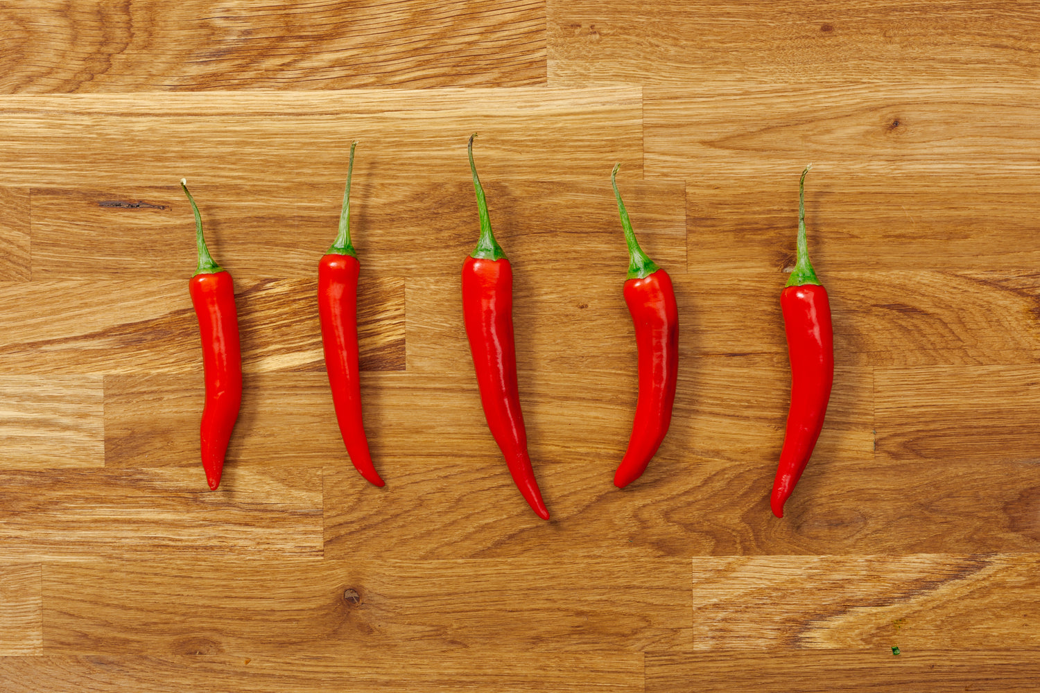 Five hot red chili peppers lined up horizontally on a wood carving board